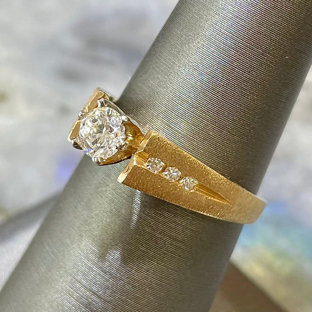 A diamond ring that features one Standard Round Brilliant Cut diamond set in a six prong head with three side diamonds channel set along side it in a 14kt yellow gold satin/matte finish mounting. The piece is on a ring holder.