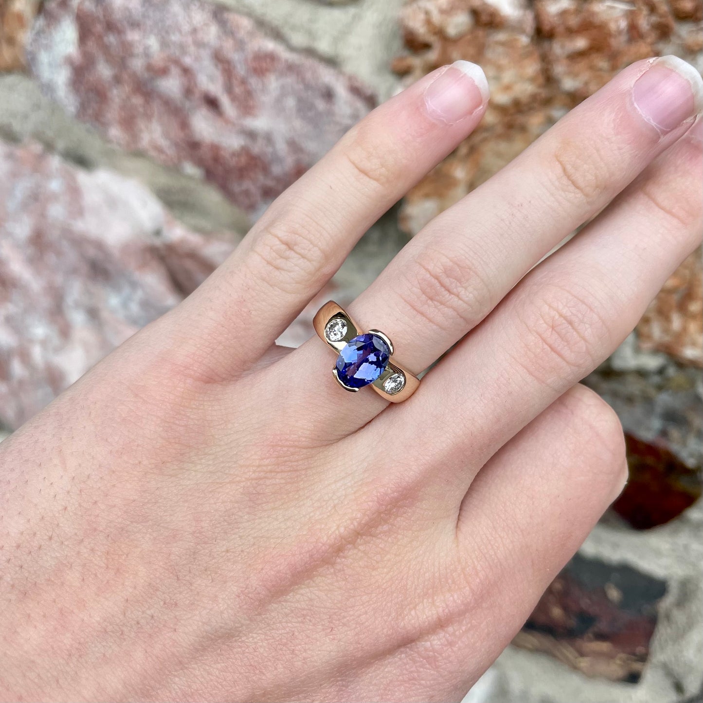 A yellow gold ring half-bezel set with an oval cut blue tanzanite and two flush set round diamonds.