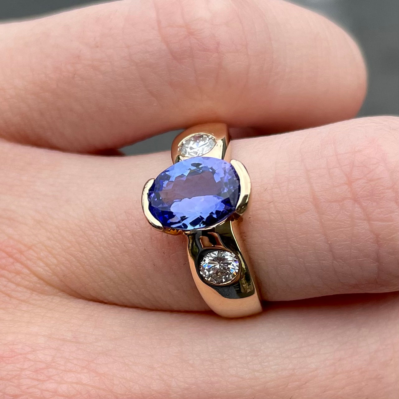 A yellow gold ring half-bezel set with an oval cut blue tanzanite and two flush set round diamonds.
