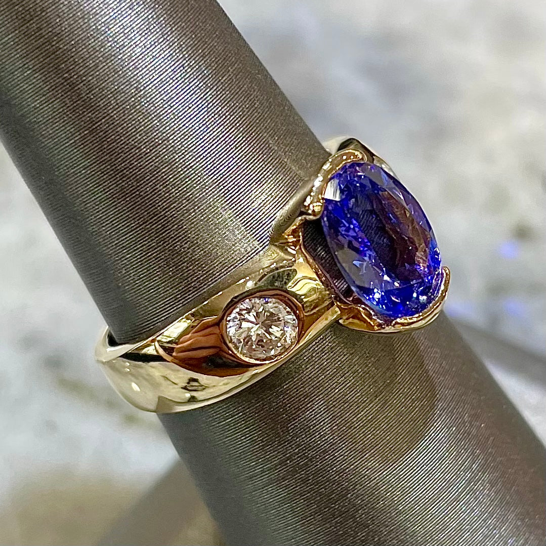 A yellow gold ring half-bezel set with an oval cut blue tanzanite and two flush set round diamonds.