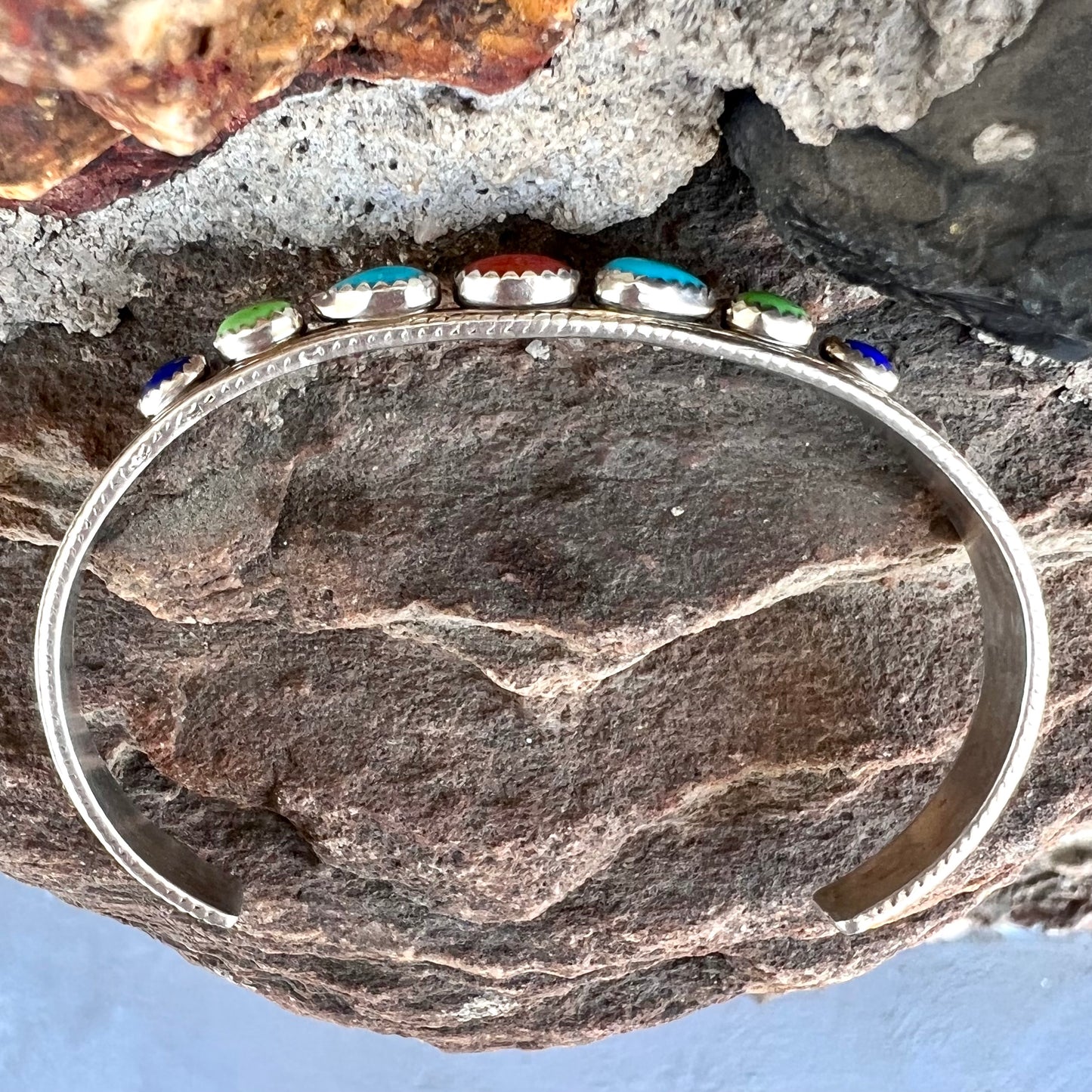 A silver rope cuff bracelet set with coral, turquoise, gaspeite, and lapis lazuli stones. The underside is stamped "Daniel Mike".