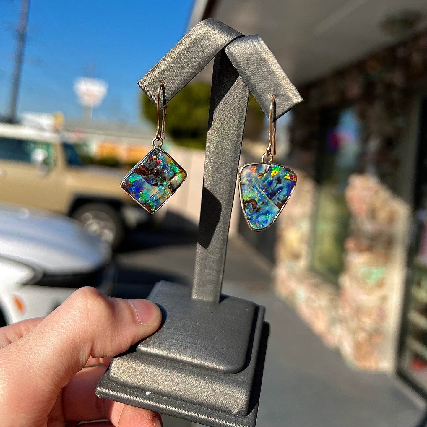 An asymmetric pair of French wire dangle yellow gold boulder opal earrings. One opal is a square, and the other is a triangle.