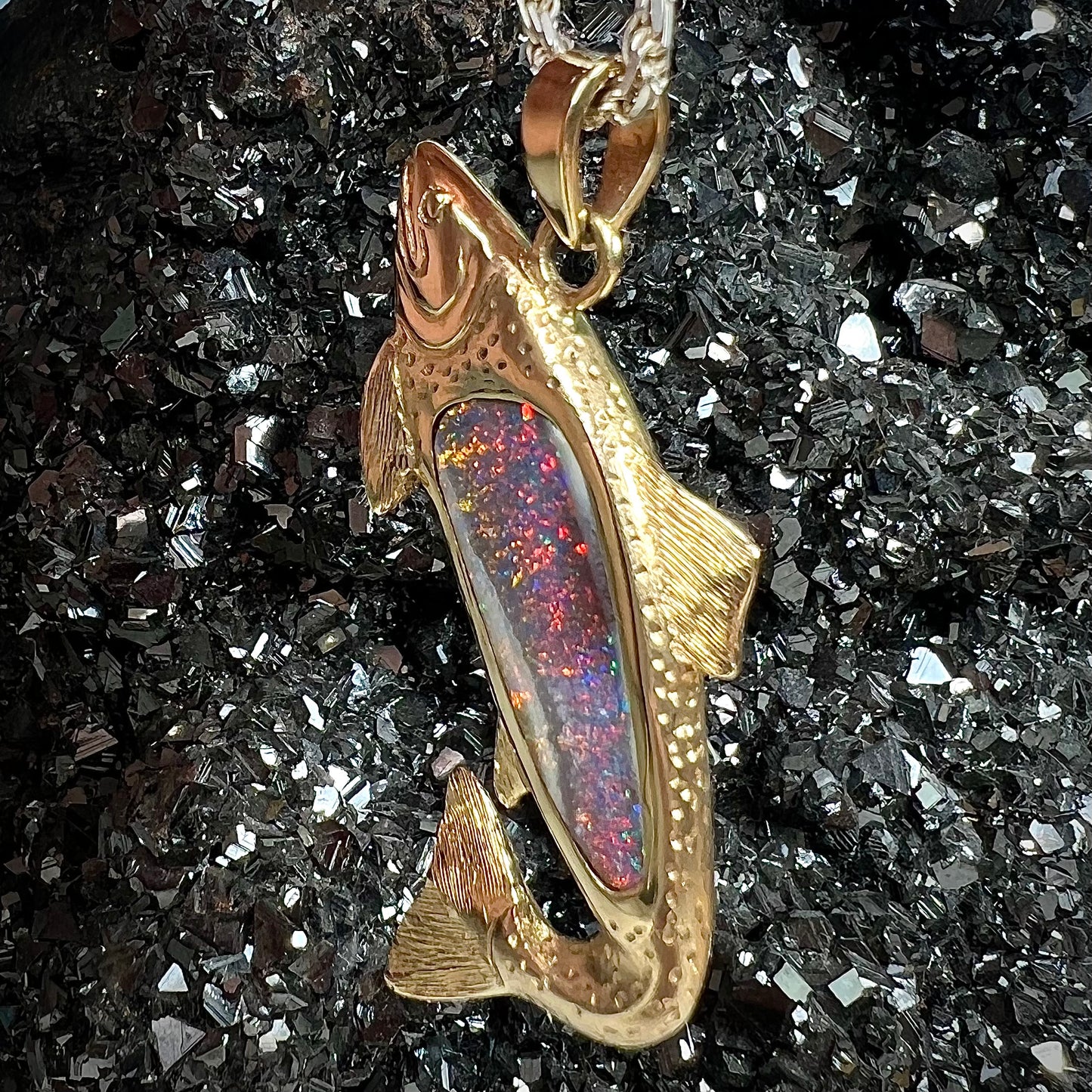 A yellow gold fish pendant detailed to look like a rainbow trout. The body of the fish is set with a color changing black opal stone.