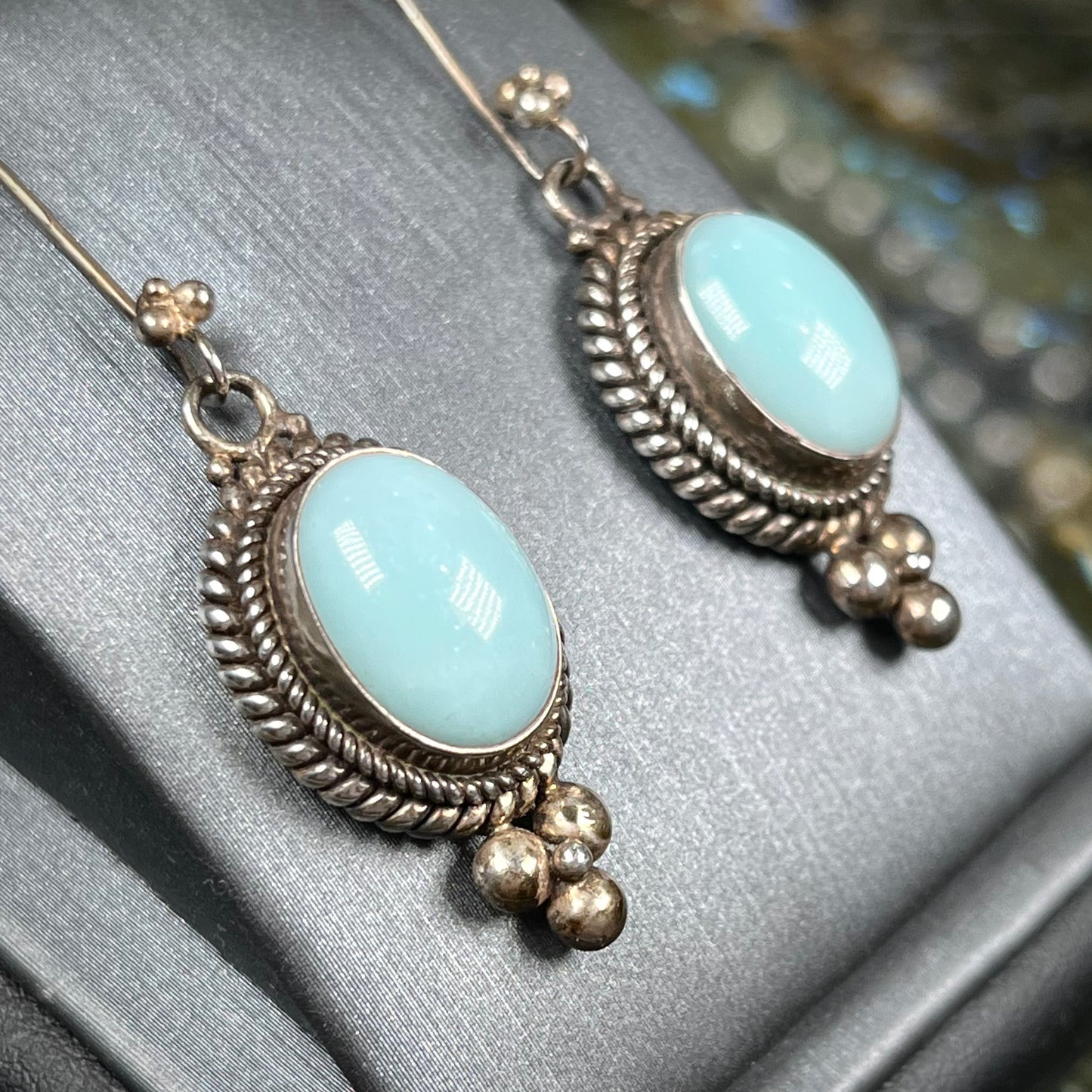 A pair of silver wire dangle earrings set with blue agate stones. The earrings have a braided rope design.