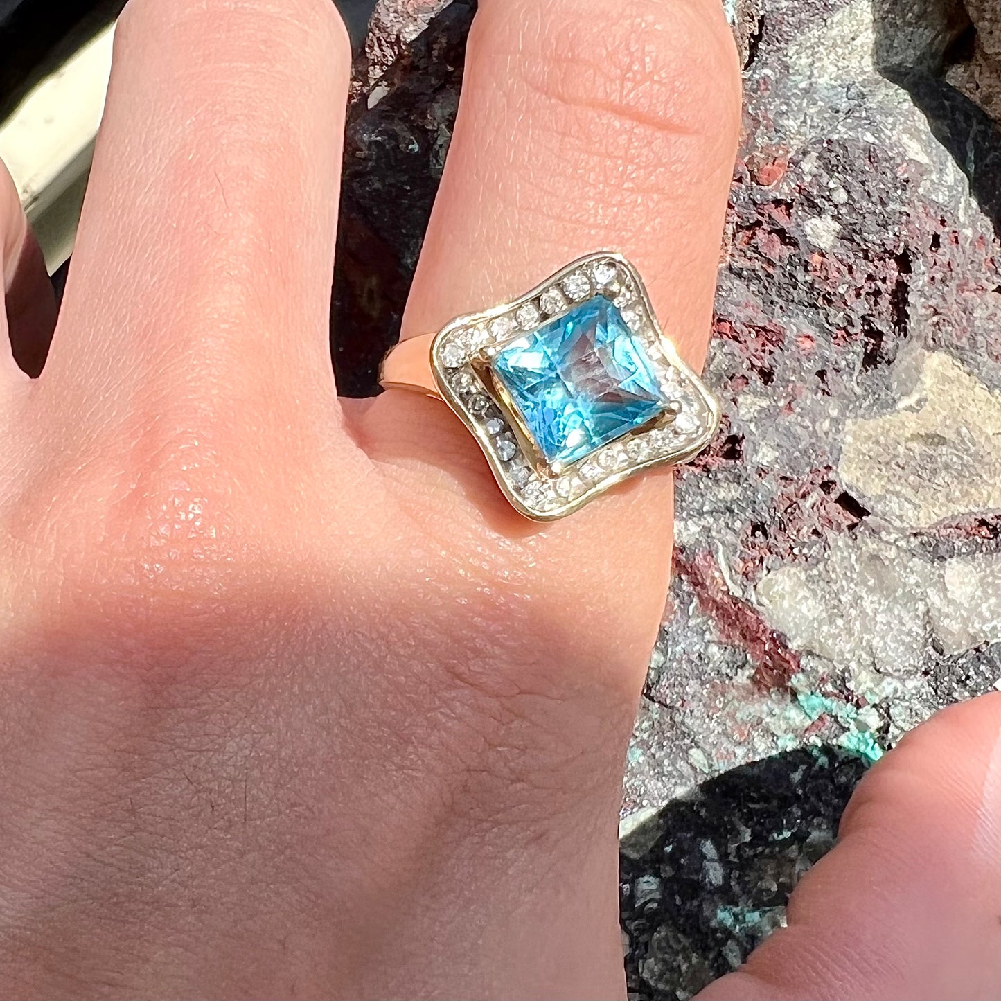 A checkerboard princess cut Swiss blue topaz ring. The stone is set in a yellow gold, diamond halo mounting.