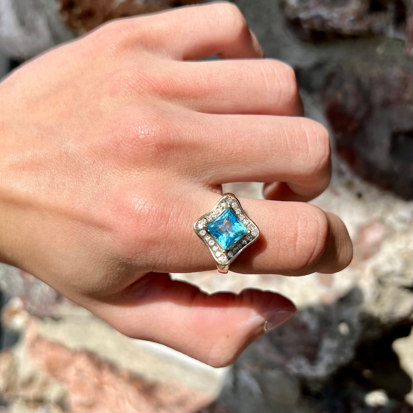 A checkerboard princess cut Swiss blue topaz ring. The stone is set in a yellow gold, diamond halo mounting.