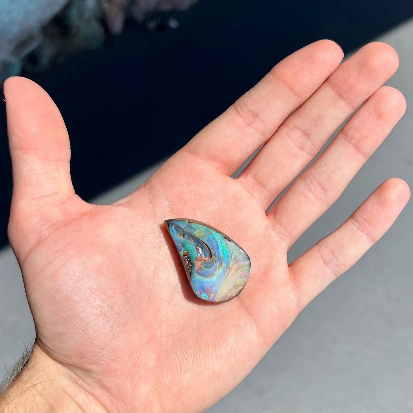 A polished pear shaped boulder opal stone from Quilpie, Australia. Predominant colors are blue, green, and aqua.