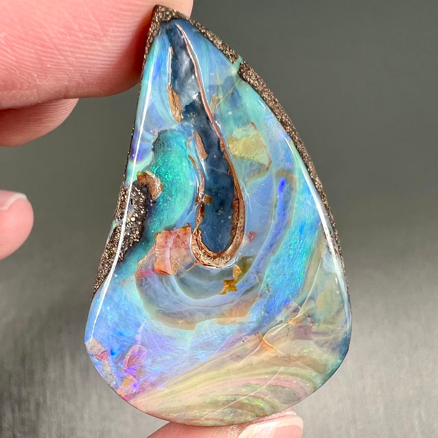 A polished pear shaped boulder opal stone from Quilpie, Australia. Predominant colors are blue, green, and aqua.