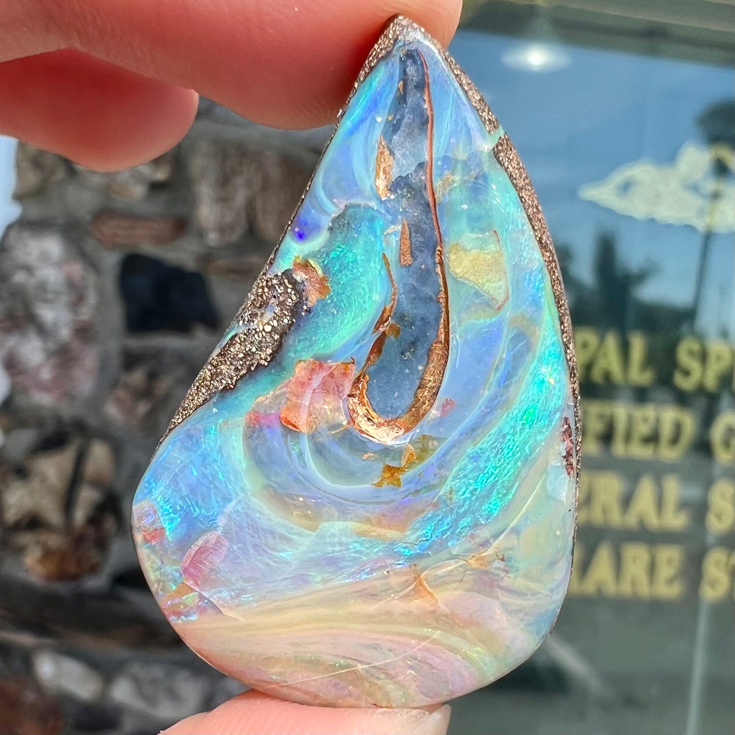 A polished pear shaped boulder opal stone from Quilpie, Australia. Predominant colors are blue, green, and aqua.