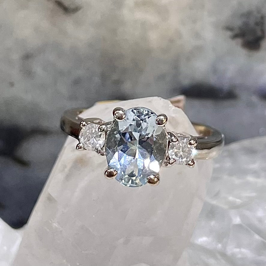 An oval cut aquamarine ring in 14kt white gold that features one diamond on each side of the center stone. The ring is resting on a white quartz crystal.