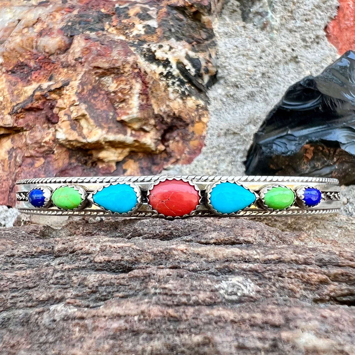 A silver rope cuff bracelet set with coral, turquoise, gaspeite, and lapis lazuli stones. The underside is stamped "Daniel Mike".
