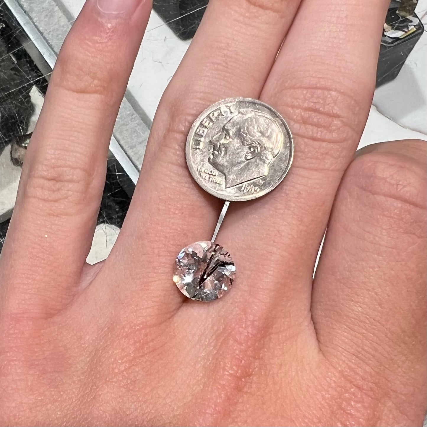 A faceted round brilliant cut clear quartz stone with black tourmaline inclusions. Inclusions appear like a clock's hands.
