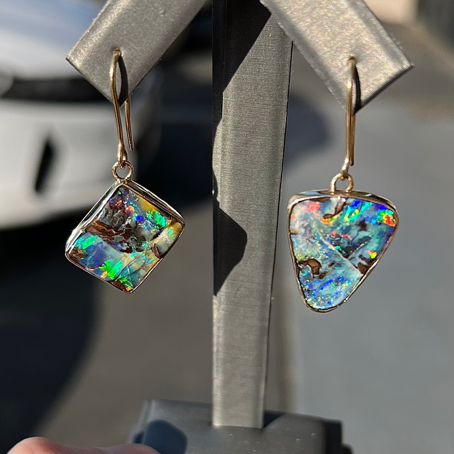 An asymmetric pair of French wire dangle yellow gold boulder opal earrings. One opal is a square, and the other is a triangle.