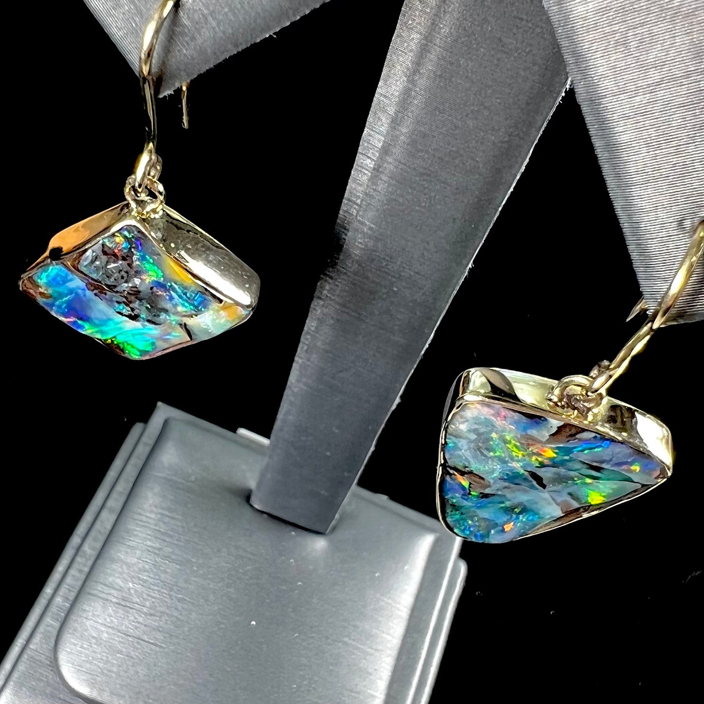 An asymmetric pair of French wire dangle yellow gold boulder opal earrings. One opal is a square, and the other is a triangle.