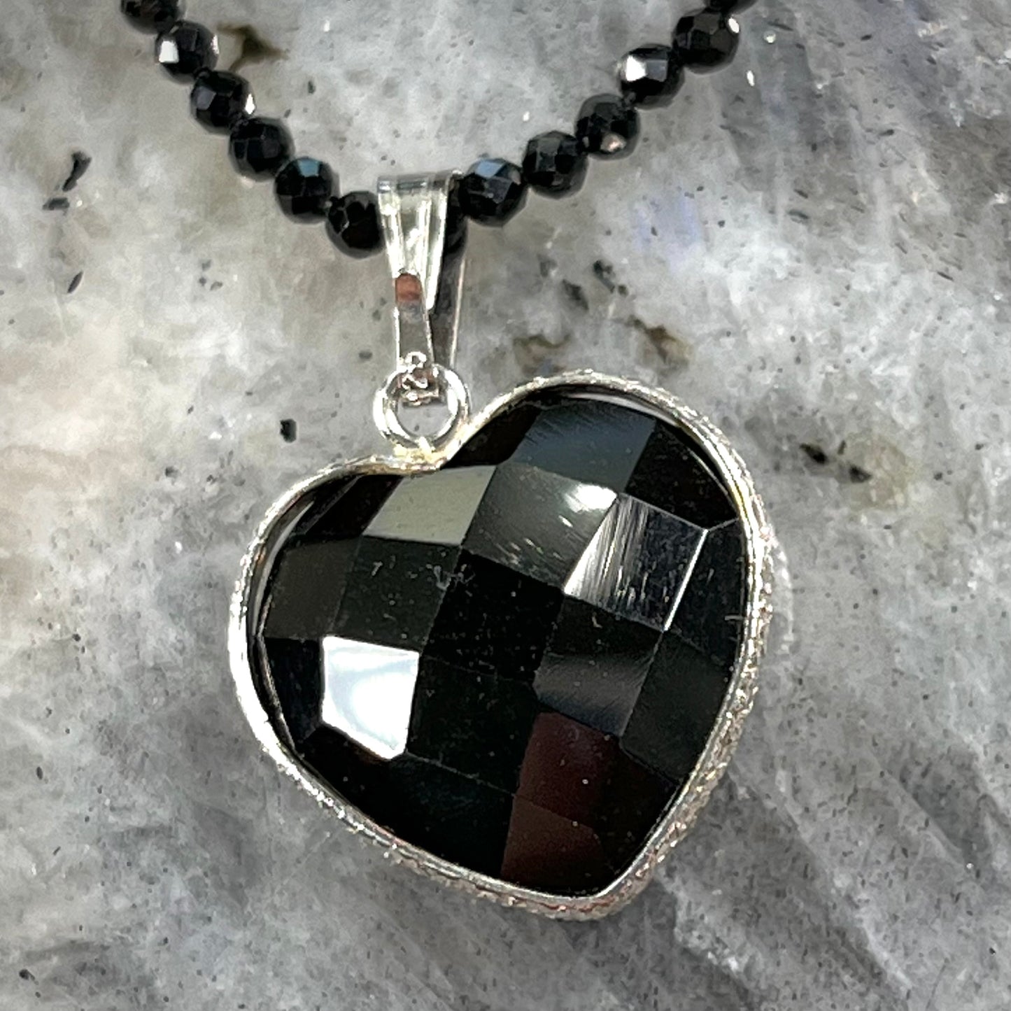 A silver heart shape black stone necklace. The heart is a faceted cut black spinel, and the pendant is on faceted black spinel beads.