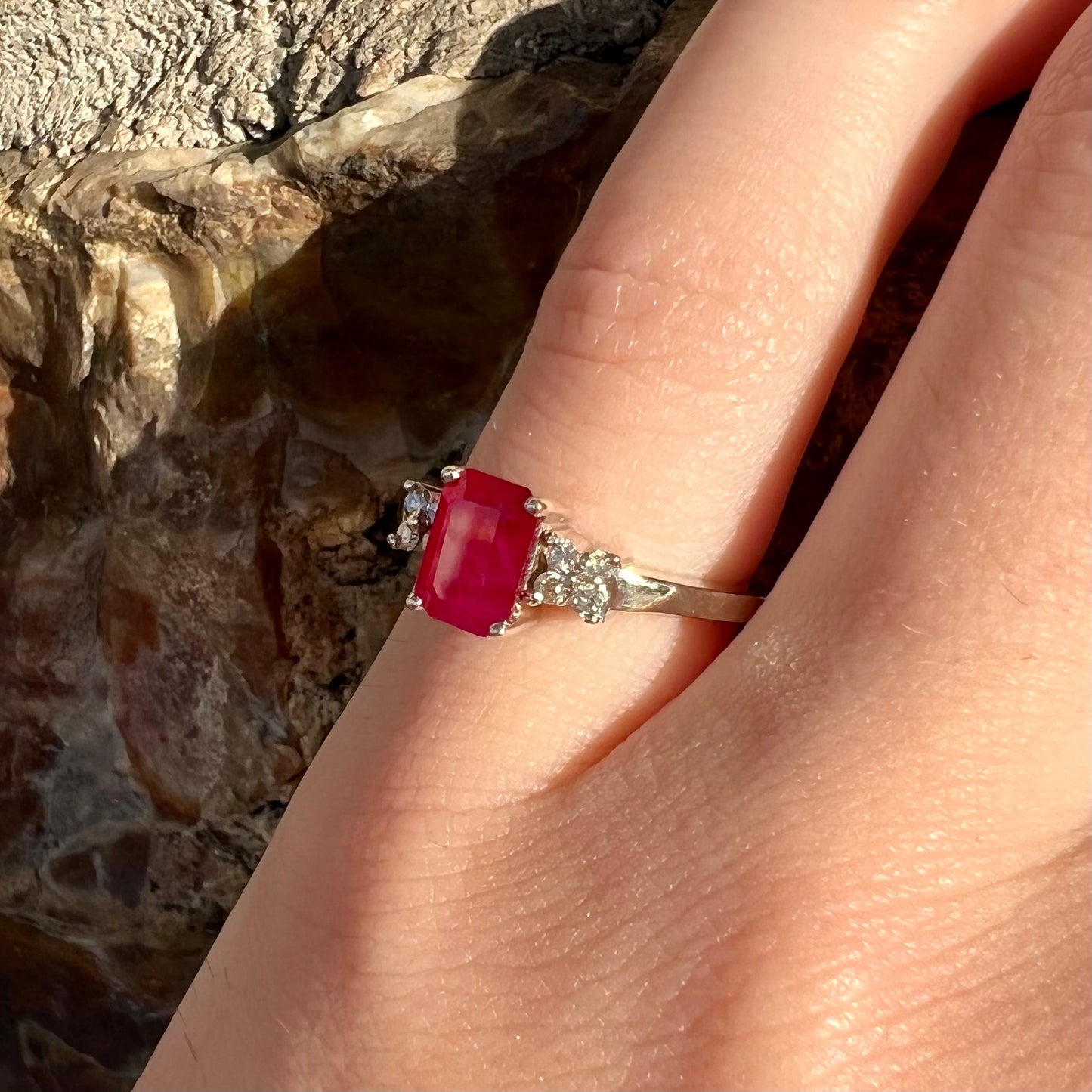 A white gold ring with an emerald cut red synthetic ruby set between four round cut diamonds on each side.