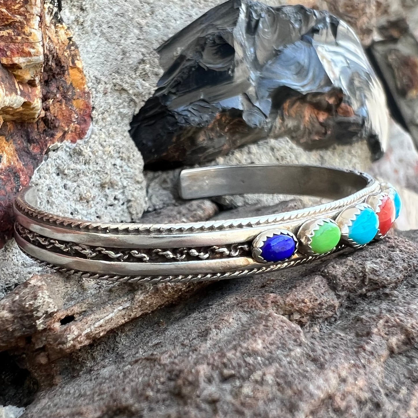 A silver rope cuff bracelet set with coral, turquoise, gaspeite, and lapis lazuli stones. The underside is stamped "Daniel Mike".