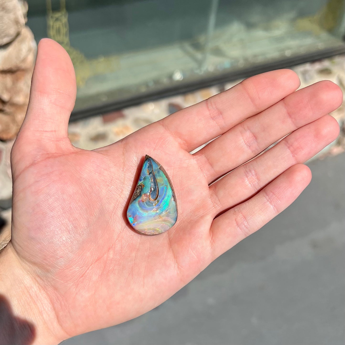 A polished pear shaped boulder opal stone from Quilpie, Australia. Predominant colors are blue, green, and aqua.