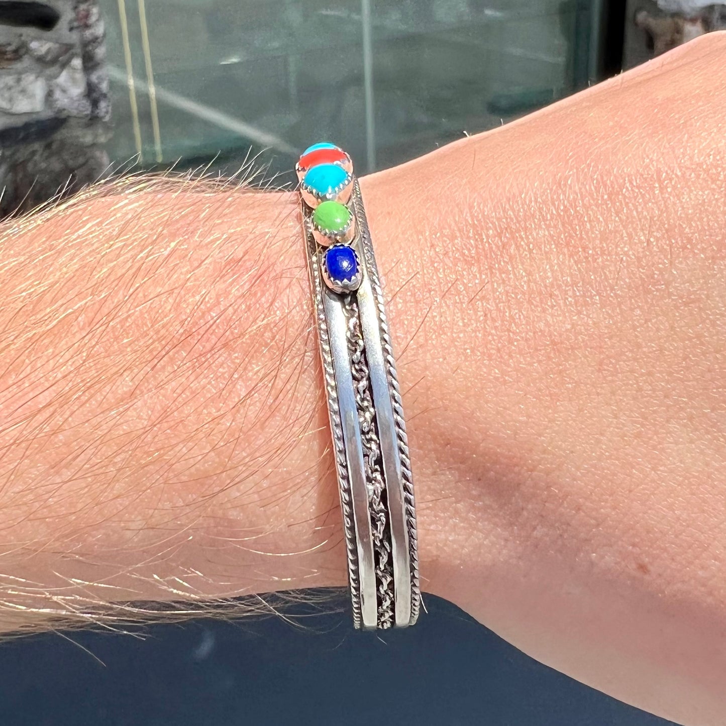 A silver rope cuff bracelet set with coral, turquoise, gaspeite, and lapis lazuli stones. The underside is stamped "Daniel Mike".