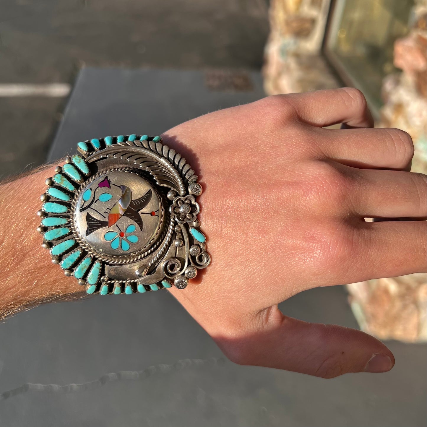 A sterling silver cuff bracelet featuring the motif of a stone inlaid hummingbird with petit point turquoise accents, handmade by Navajo artist Leo G. Harvey.
