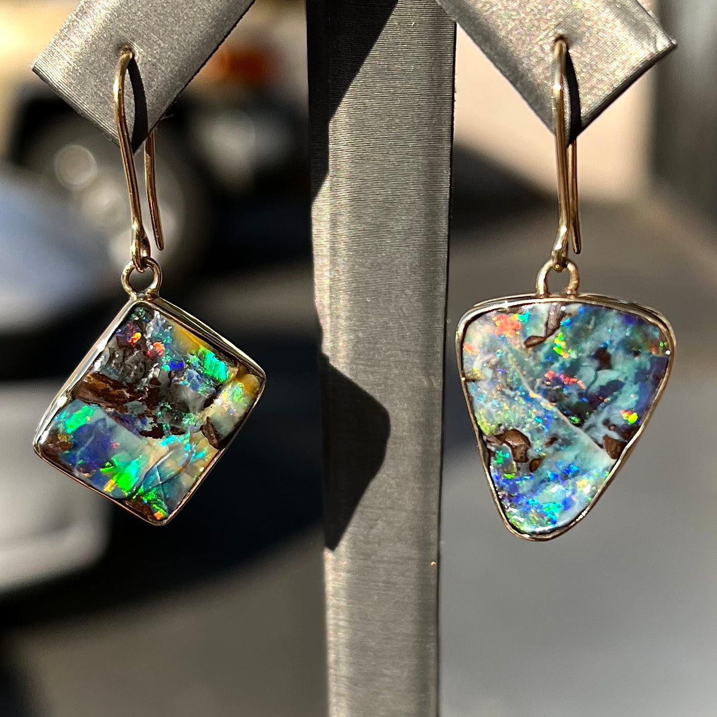An asymmetric pair of French wire dangle yellow gold boulder opal earrings. One opal is a square, and the other is a triangle.