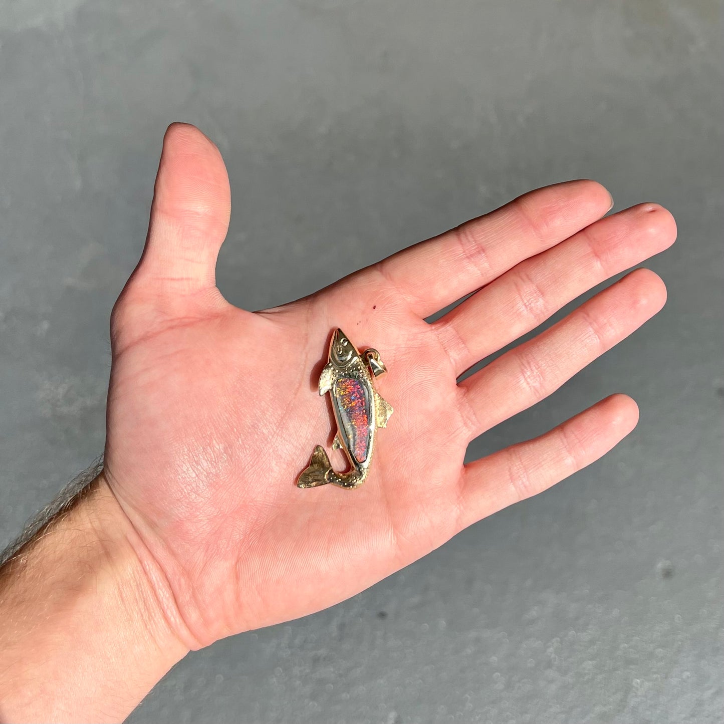 A yellow gold fish pendant detailed to look like a rainbow trout. The body of the fish is set with a color changing black opal stone.
