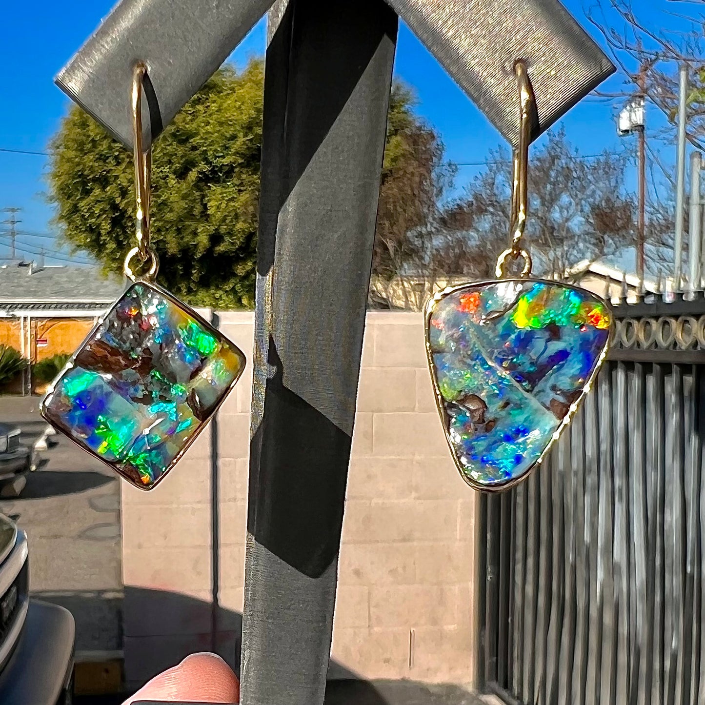 An asymmetric pair of French wire dangle yellow gold boulder opal earrings. One opal is a square, and the other is a triangle.