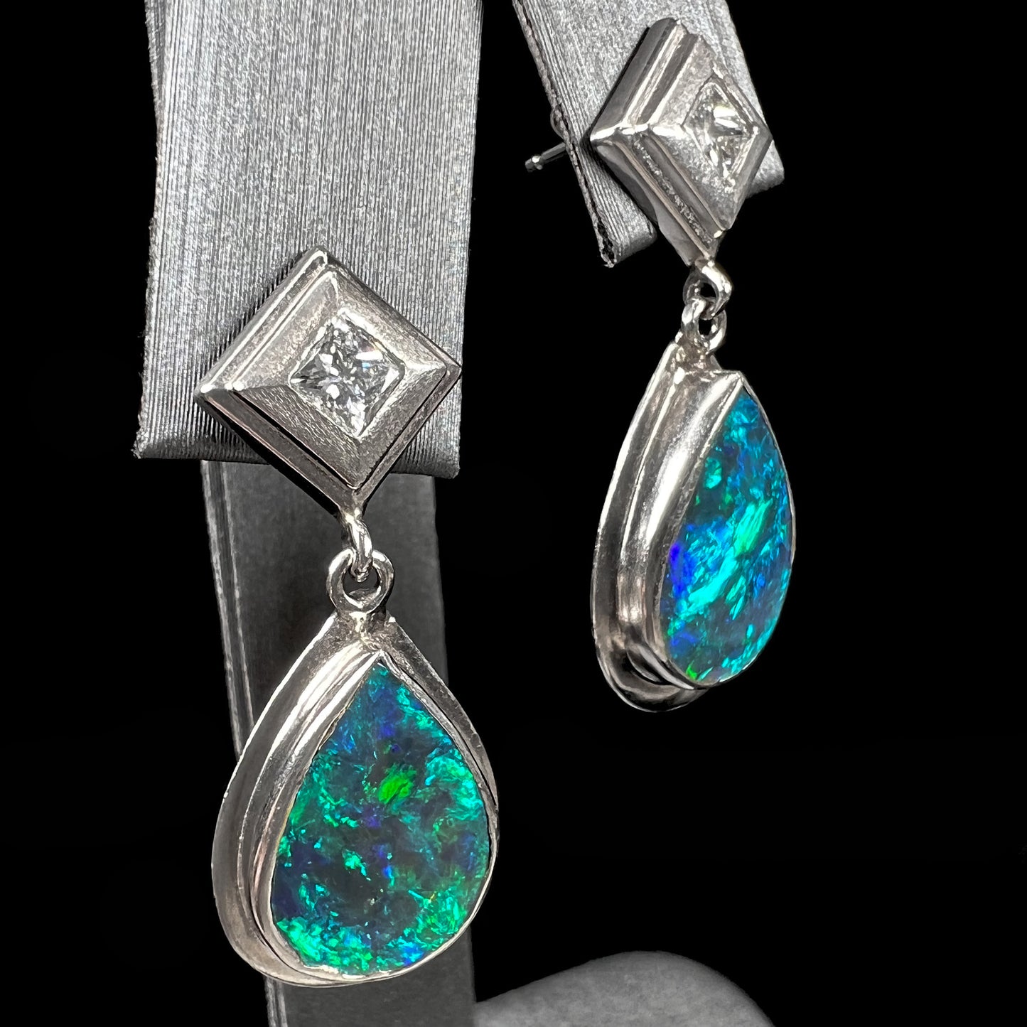 Black crystal opal dangle earrings with blue body color and green play of color set with princess cut diamonds in platinum.