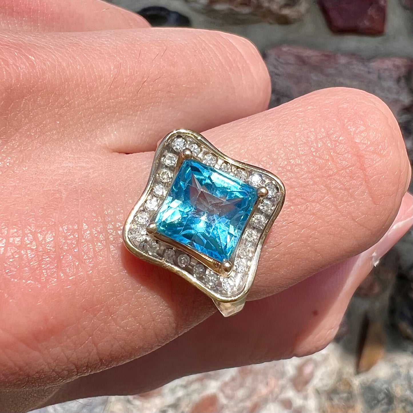 A checkerboard princess cut Swiss blue topaz ring. The stone is set in a yellow gold, diamond halo mounting.