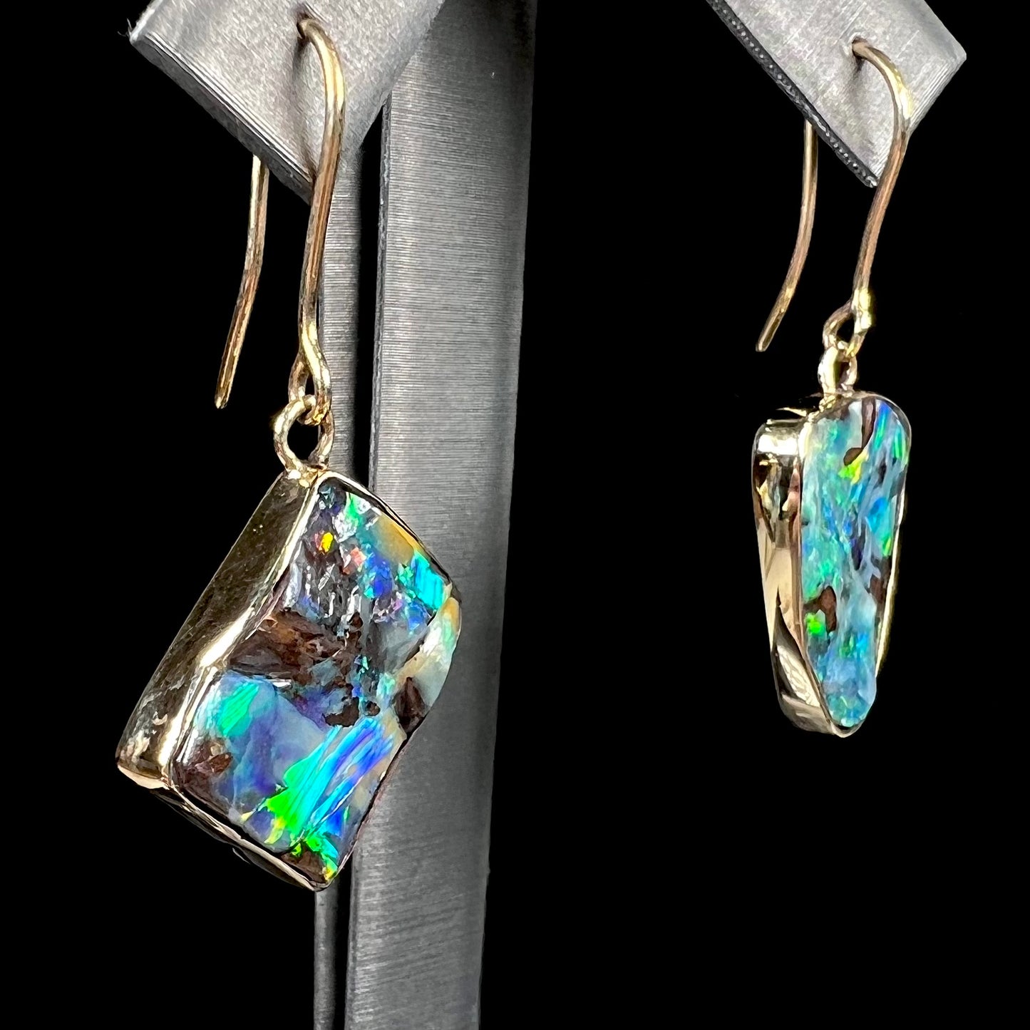 An asymmetric pair of French wire dangle yellow gold boulder opal earrings. One opal is a square, and the other is a triangle.