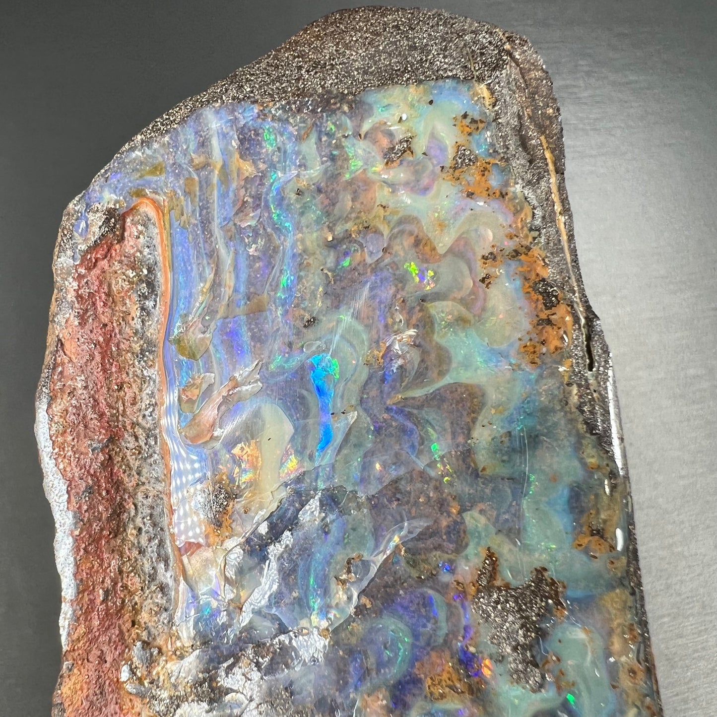 A 7 inch long, polished Quilpie boulder opal specimen. The stone shows colors of blue, green, and orange.