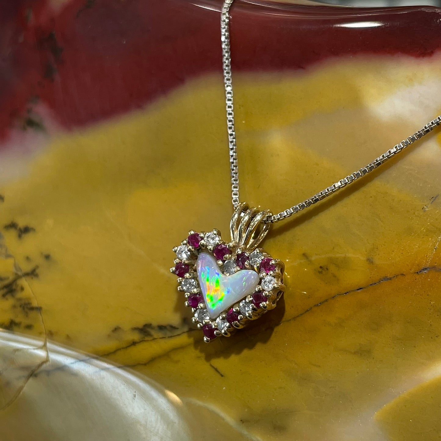 A yellow gold pendant set with a heart cut white opal in a halo of alternating round rubies and diamonds.