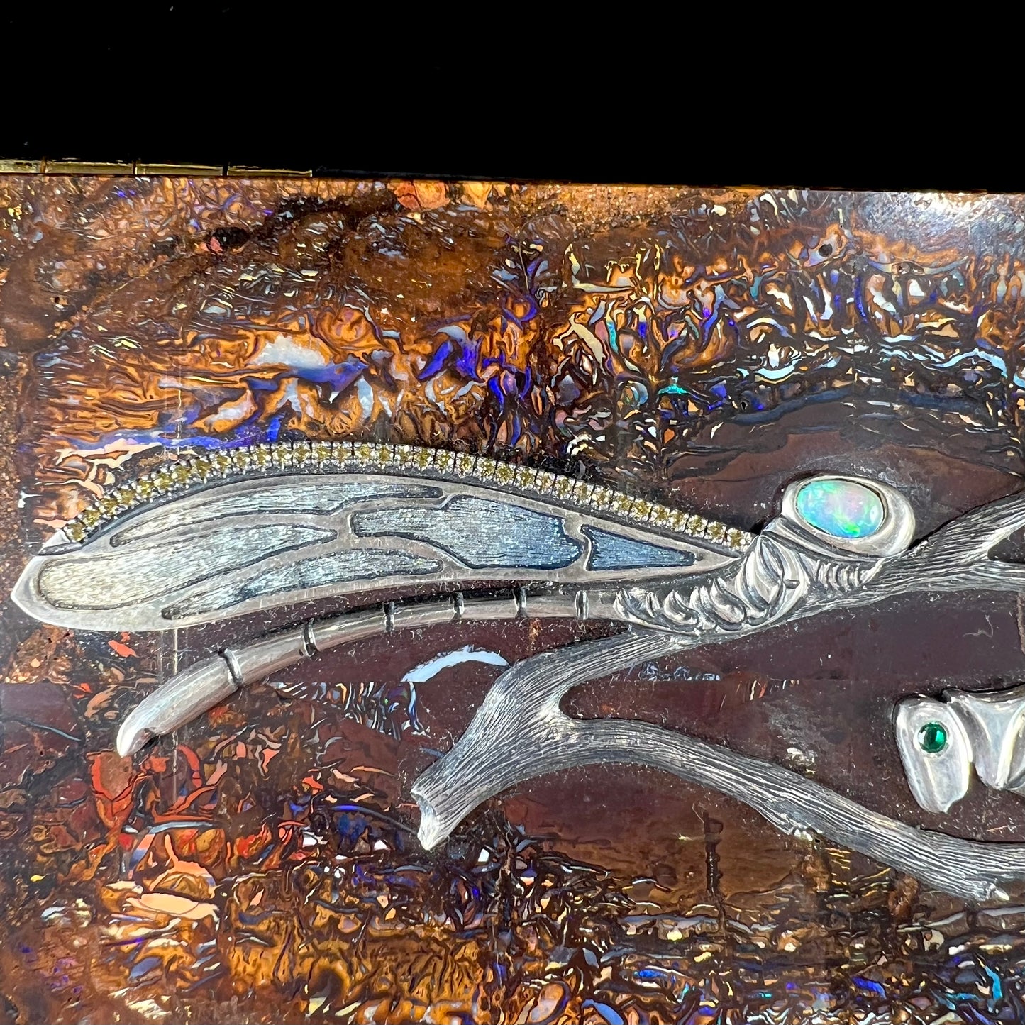 A stone box made from Koroit, Australian boulder opal featuring the motif of a dragonfly and cricket made from sterling silver, set with yellow diamonds, an emerald, and a crystal opal.