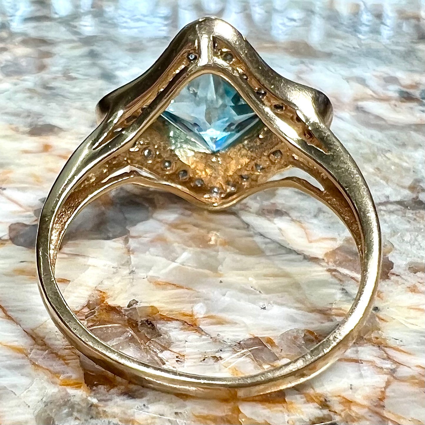 A checkerboard princess cut Swiss blue topaz ring. The stone is set in a yellow gold, diamond halo mounting.