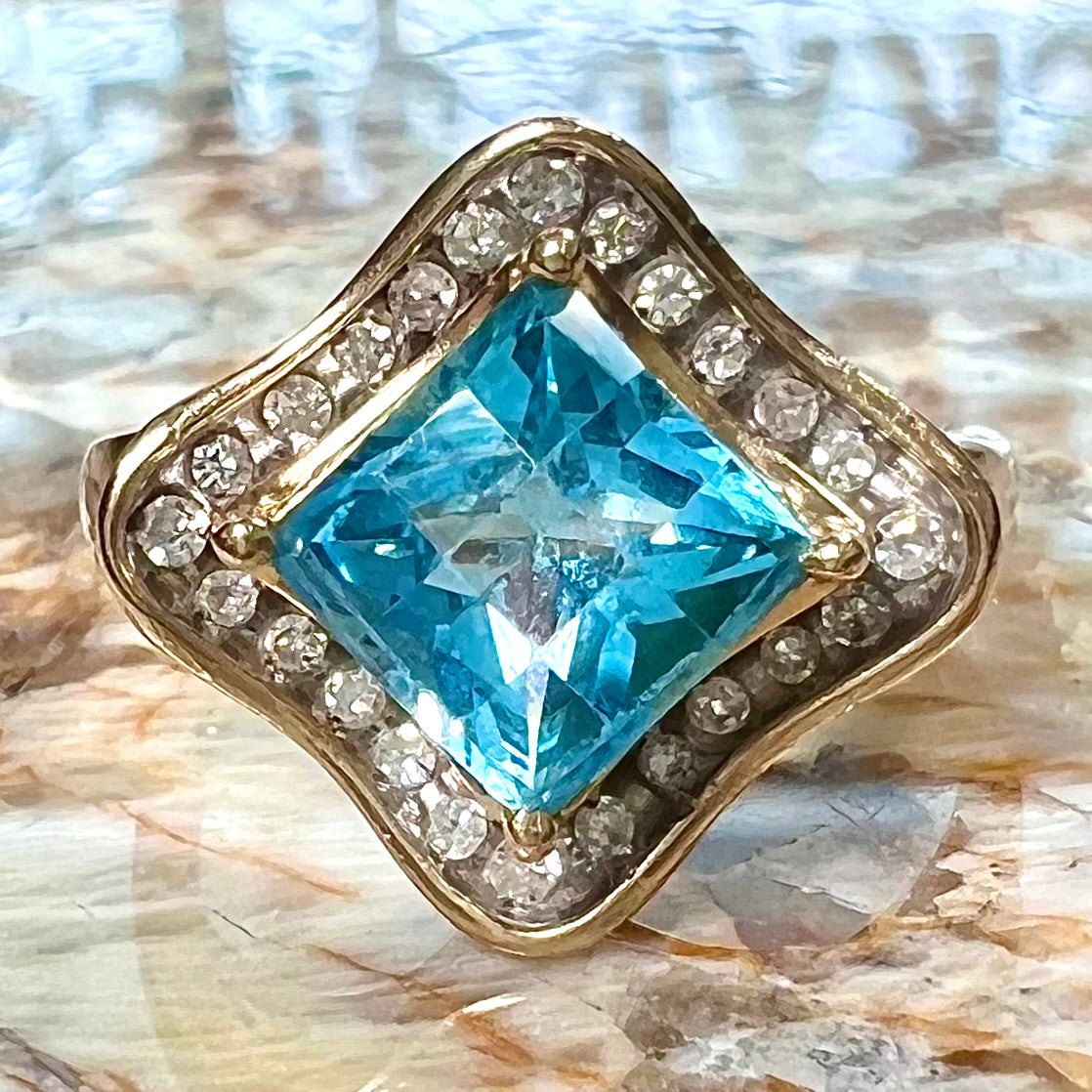 A checkerboard princess cut Swiss blue topaz ring. The stone is set in a yellow gold, diamond halo mounting.
