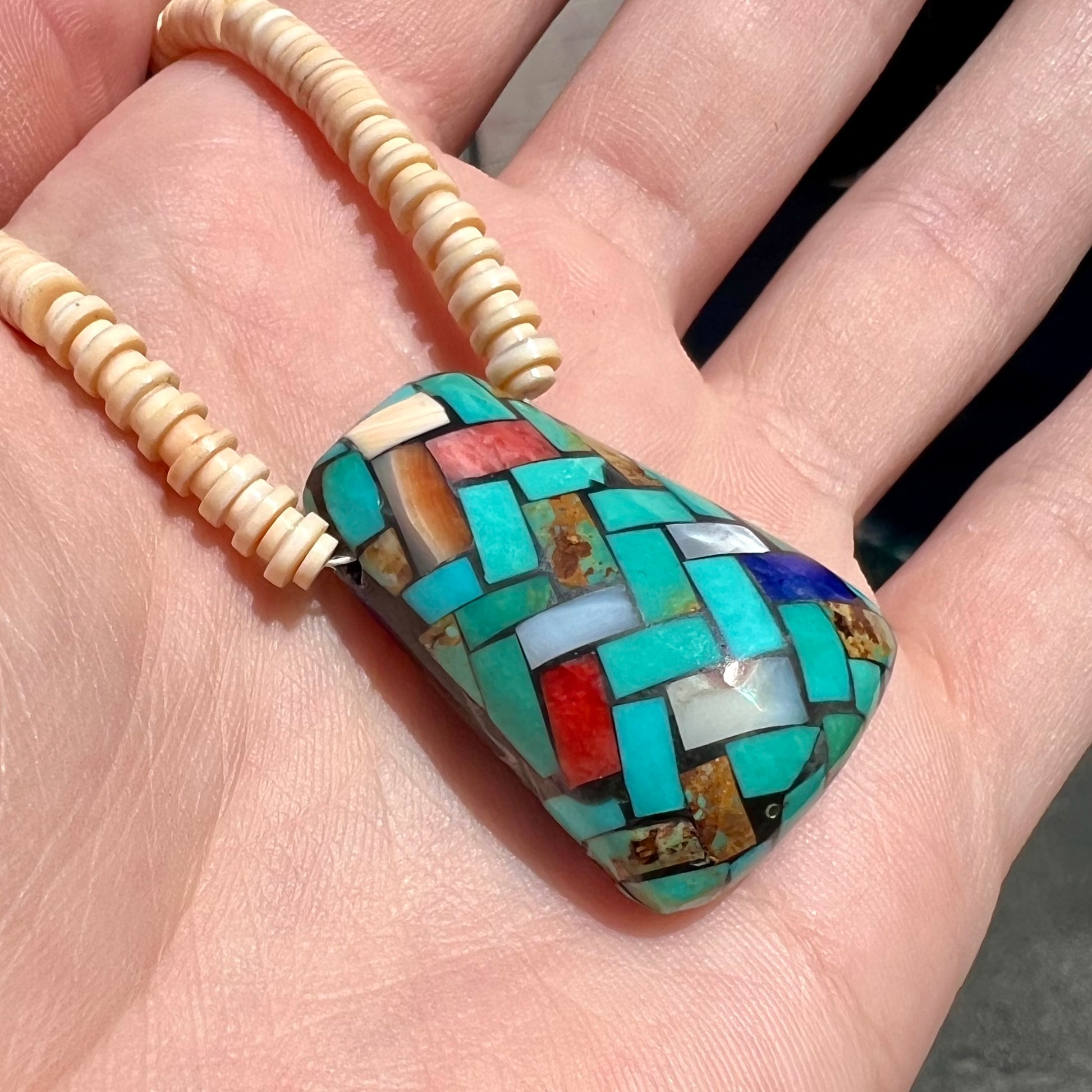 A three dimensional mosaic stone inlay pendant set with turquoise, lapis lazuli, coral, and mother of pearl on a puka shell necklace.