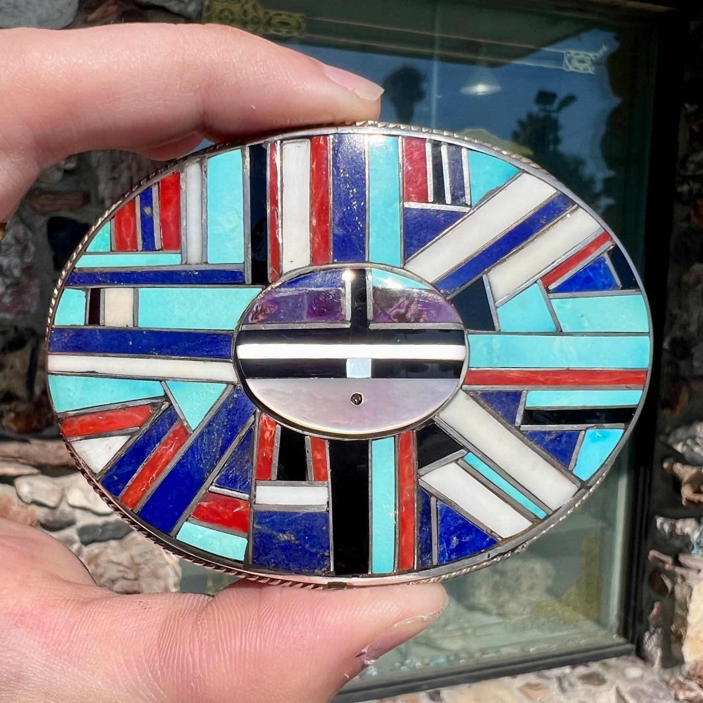 A sterling silver Zuni sunface belt buckle inlaid with lapis, turquoise, onyx, mother of pearl, coral, and sugilite stones. It is stamped "RGB Zuni."