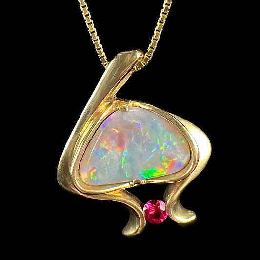 A ladies' yellow gold pendant bezel set with a natural black opal and pink sapphire accent.  The opal shines red, green, blue, and purple.