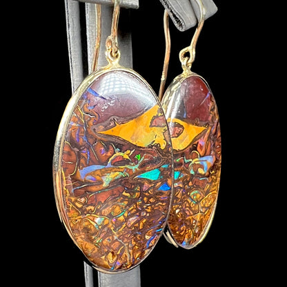 A pair of oval cut Koroit boulder opal dangle earrings in yellow gold bezel frames.  The opal exhibits a unique pattern and blue colors.