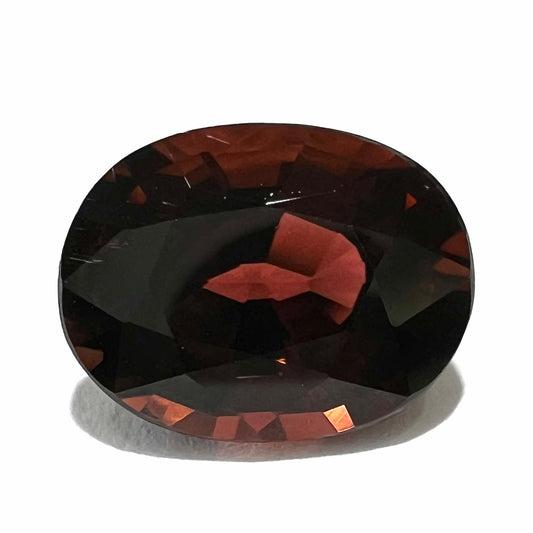 A loose, faceted oval cut dravite tourmaline gemstone.  The stone is a dark brown color with a purple secondary color.