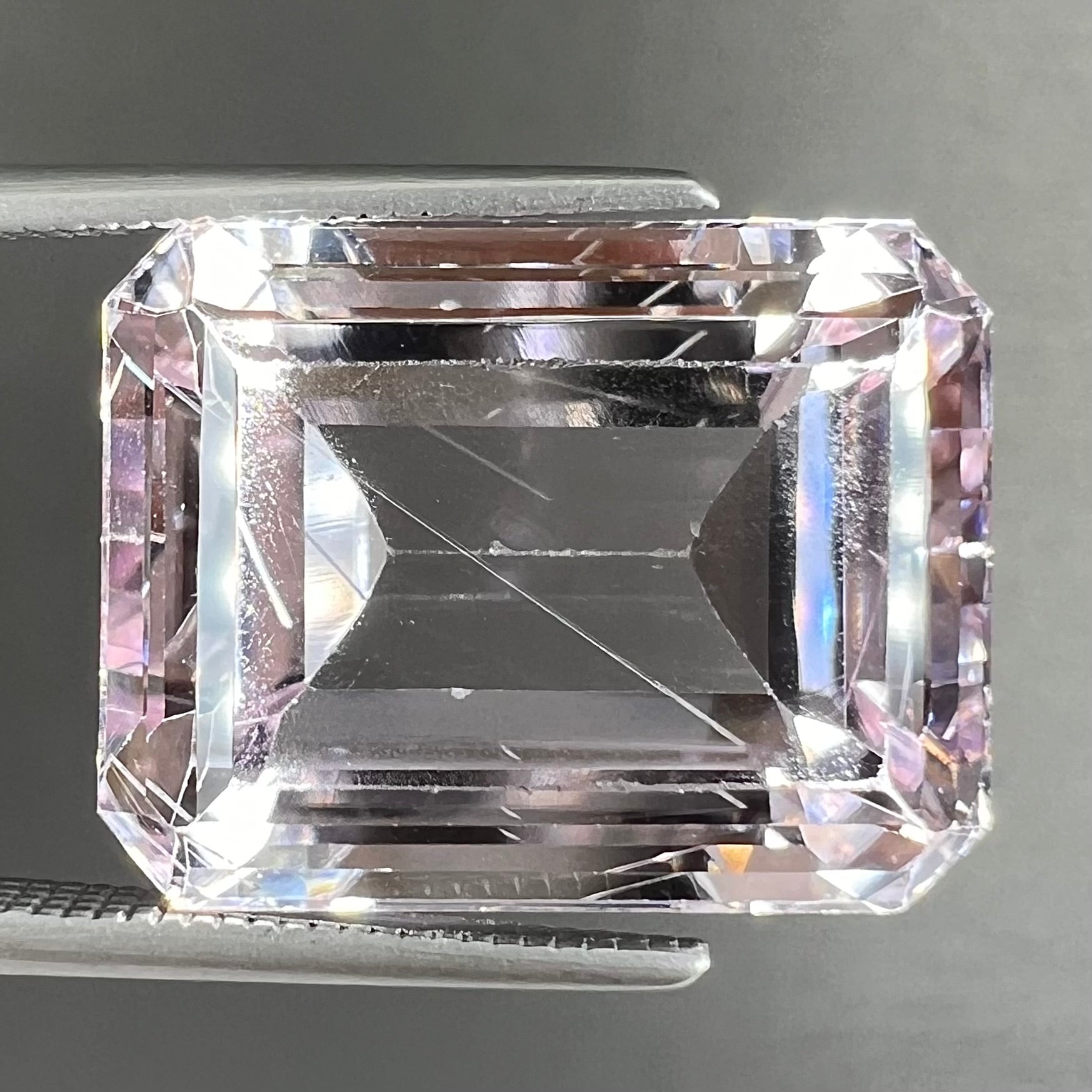 A loose, emerald cut kunzite gemstone.  The stone has eye-visible rutile needle inclusions and the table is abraded.
