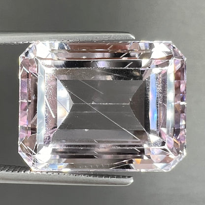 A loose, emerald cut kunzite gemstone.  The stone has eye-visible rutile needle inclusions and the table is abraded.