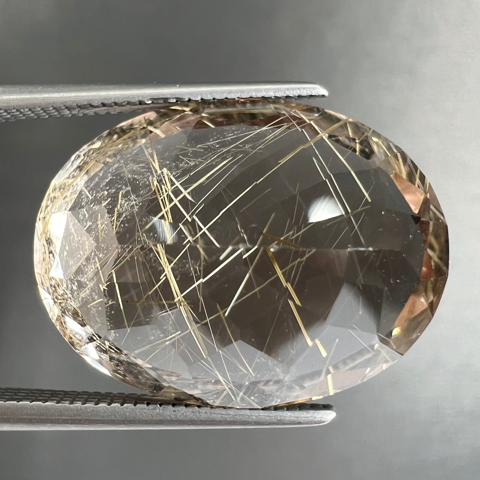 A faceted oval cut rutilated quartz gemstone.  The stone is smoky color with golden rutile needle inclusions.