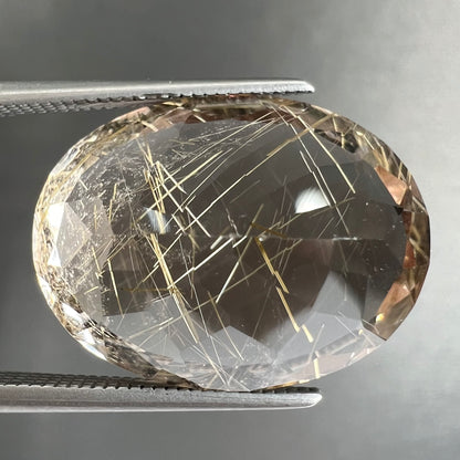 A faceted oval cut rutilated quartz gemstone.  The stone is smoky color with golden rutile needle inclusions.