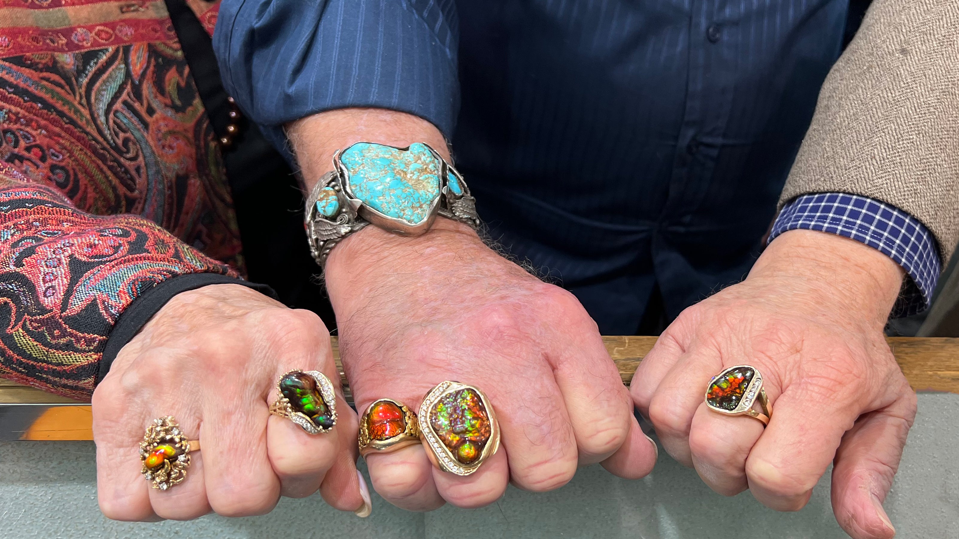 Three peoples' hands, one woman and two men, wearing gold rings set with vibrantly colorful fire agate stones.