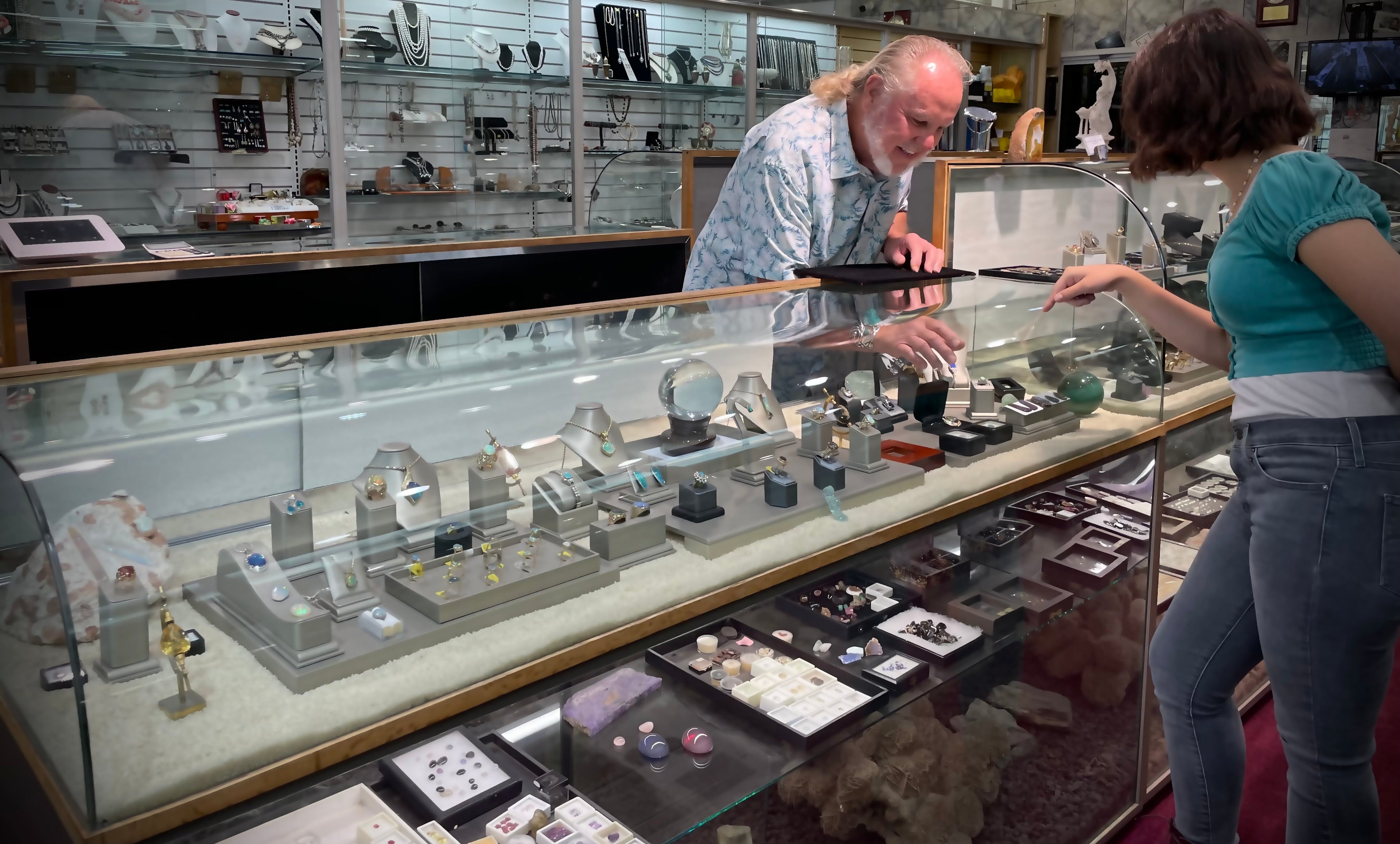 A customer is helped by a smiling jeweler.  Many gems, stones, and rings are seen.