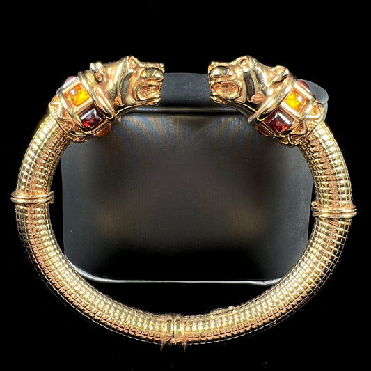 A 14 karat yellow gold hinged bangle bracelet featuring the motif of two panther heads.  There are yellow and red accent stones.