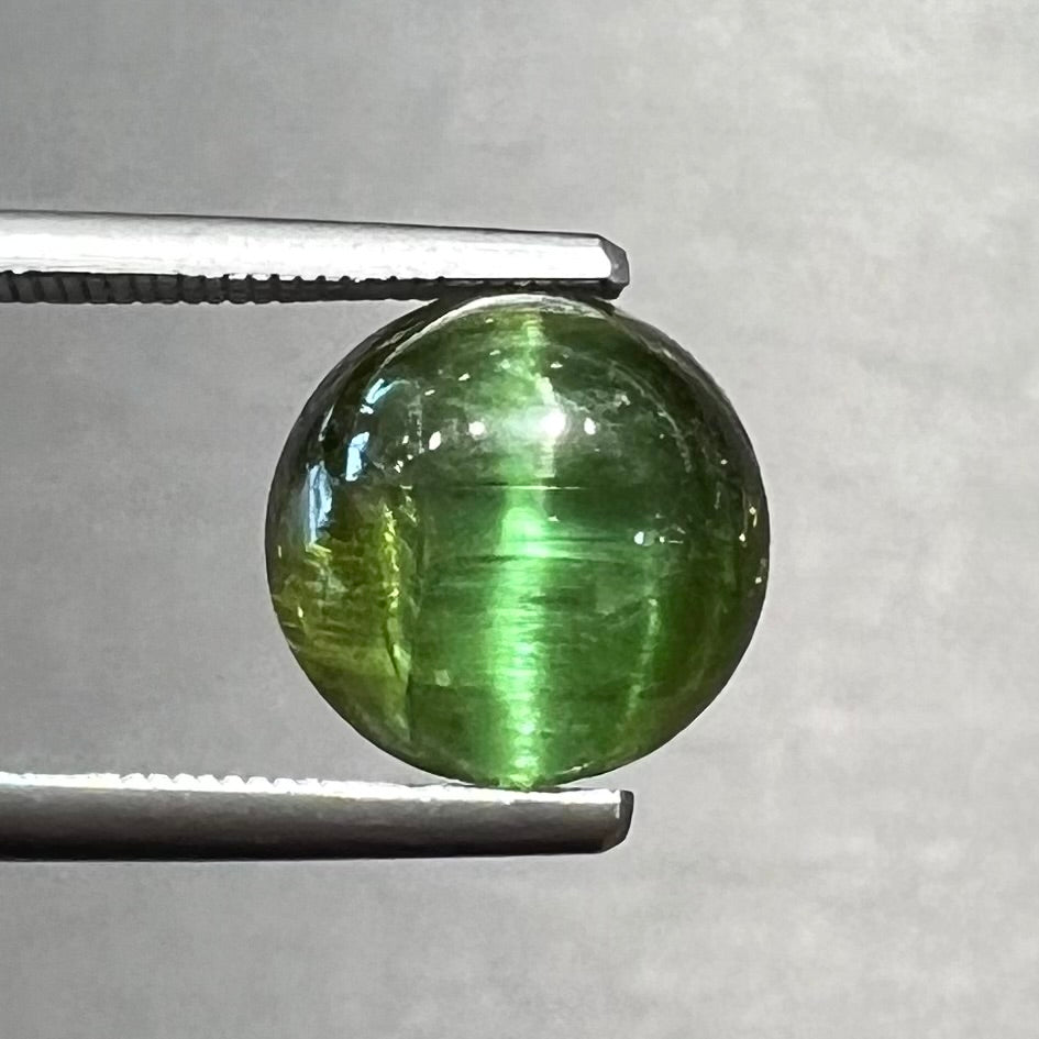A loose, round cabochon cut green tourmaline stone that displays a cat's eye phenomenon.