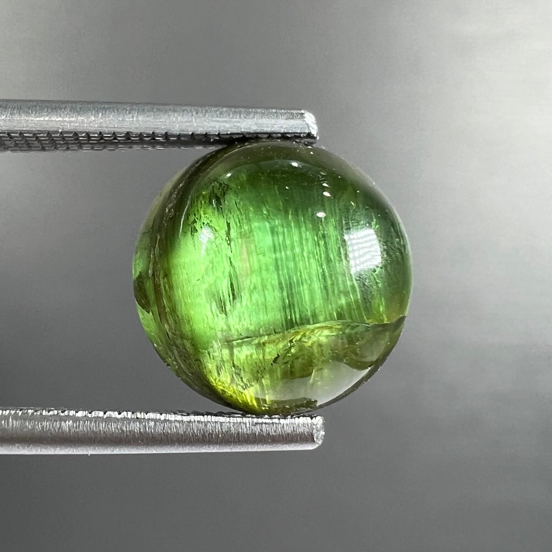 A loose, round cabochon cut green tourmaline stone that displays a cat's eye phenomenon.