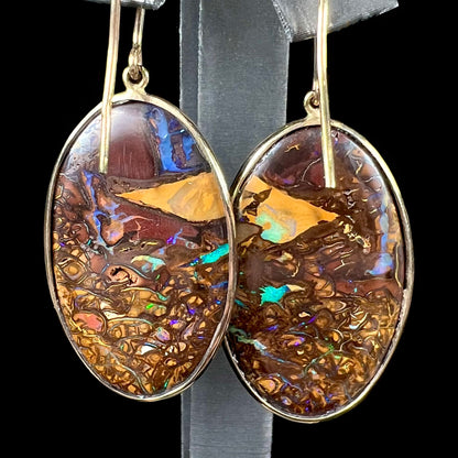 A pair of oval cut Koroit boulder opal dangle earrings in yellow gold bezel frames.  The opal exhibits a unique pattern and blue colors.